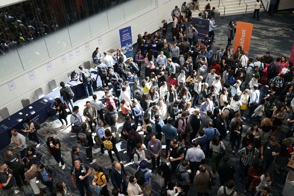 A dense crowd of people at a conference, with various booths and banners set up in the background.